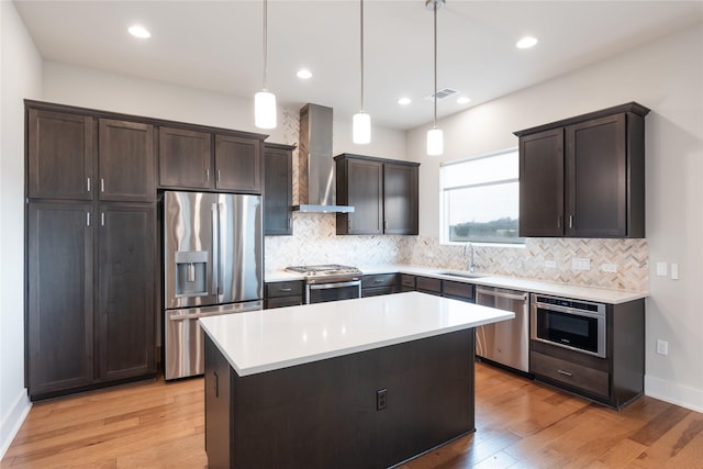 kitchen featuring wall chimney exhaust hood, a kitchen island, appliances with stainless steel finishes, decorative light fixtures, and light hardwood / wood-style floors