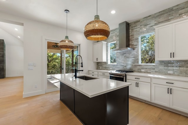 kitchen featuring sink, stainless steel range with gas stovetop, an island with sink, wall chimney range hood, and white cabinets