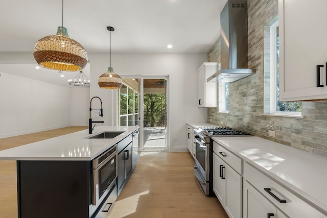 kitchen featuring sink, appliances with stainless steel finishes, wall chimney range hood, a kitchen island with sink, and white cabinets