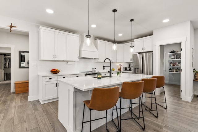 kitchen featuring custom exhaust hood, a center island with sink, stainless steel fridge with ice dispenser, white cabinetry, and hanging light fixtures