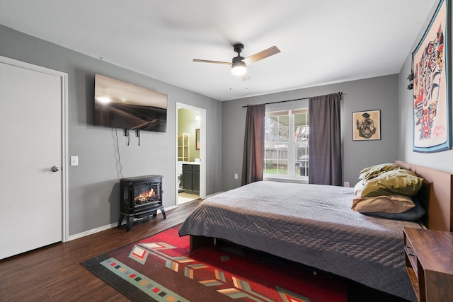 bedroom with connected bathroom, ceiling fan, a wood stove, and dark wood-type flooring