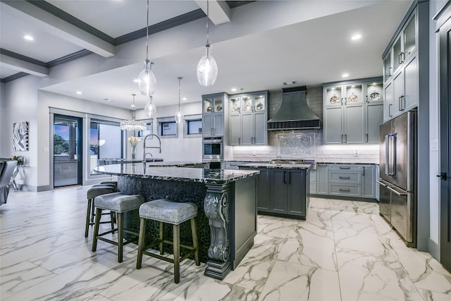 kitchen with stainless steel appliances, custom range hood, decorative light fixtures, and a kitchen island with sink