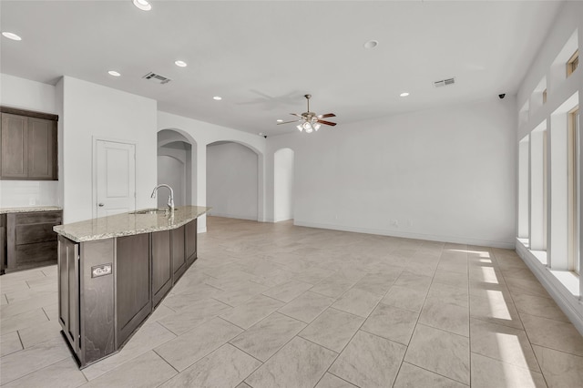 kitchen featuring ceiling fan, dark brown cabinets, light stone counters, and an island with sink