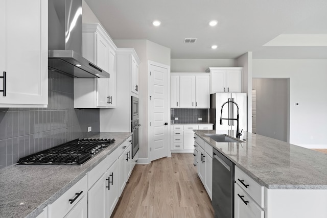 kitchen with stainless steel appliances, sink, white cabinetry, wall chimney range hood, and a kitchen island with sink