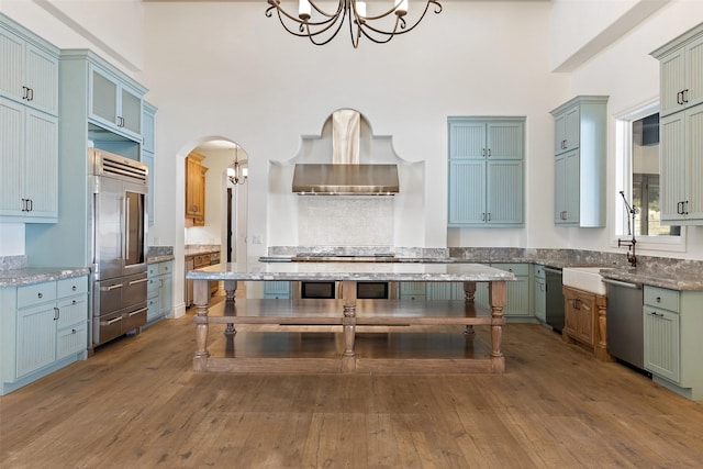 kitchen featuring hardwood / wood-style floors, light stone countertops, wall chimney range hood, pendant lighting, and appliances with stainless steel finishes