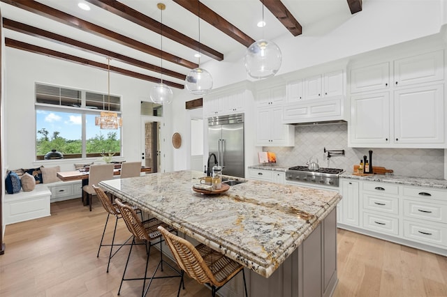 kitchen featuring built in fridge, beamed ceiling, hanging light fixtures, light stone countertops, and a center island with sink