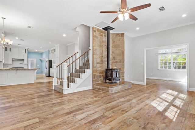 unfurnished living room featuring ceiling fan with notable chandelier, a wood stove, crown molding, and light hardwood / wood-style flooring