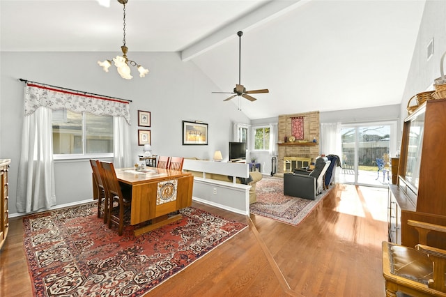 dining space with a brick fireplace, high vaulted ceiling, ceiling fan with notable chandelier, wood-type flooring, and beamed ceiling