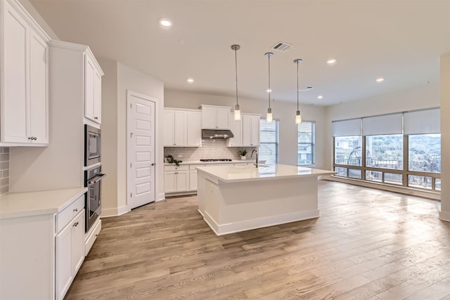 kitchen with light hardwood / wood-style flooring, hanging light fixtures, an island with sink, white cabinets, and appliances with stainless steel finishes