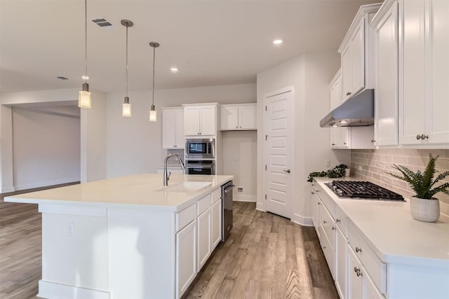 kitchen with white cabinets, an island with sink, hanging light fixtures, light countertops, and under cabinet range hood