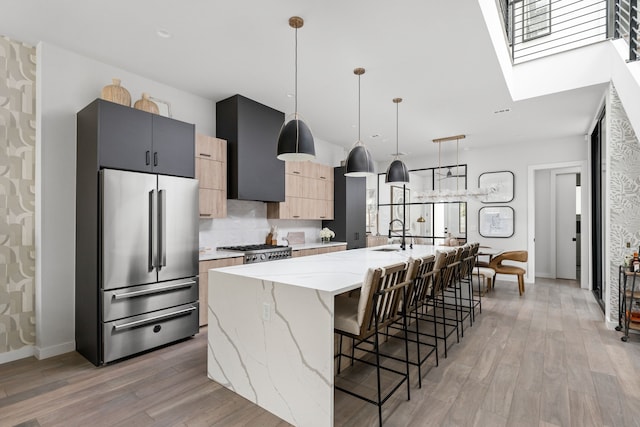 kitchen featuring an island with sink, stainless steel refrigerator, light brown cabinetry, hanging light fixtures, and light stone countertops