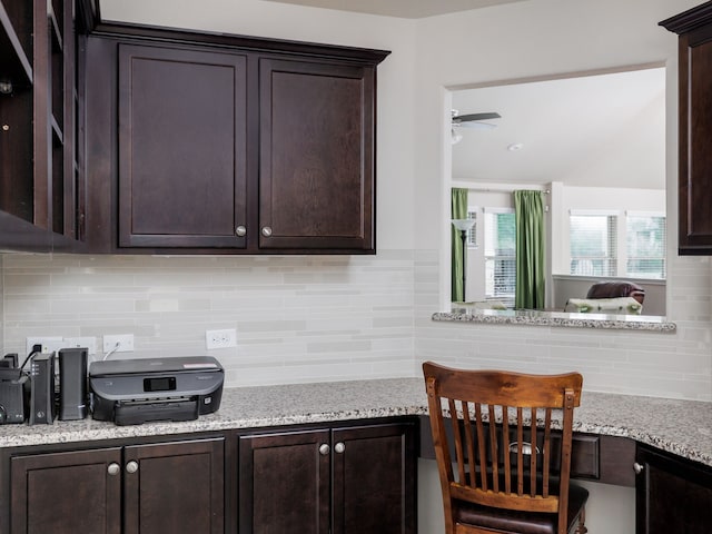 kitchen with built in desk, light stone countertops, ceiling fan, dark brown cabinetry, and tasteful backsplash