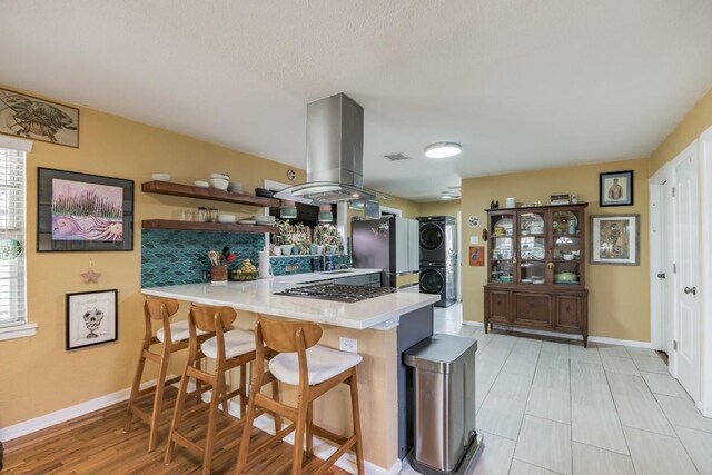 kitchen with open shelves, stacked washing maching and dryer, wall oven, a peninsula, and island range hood