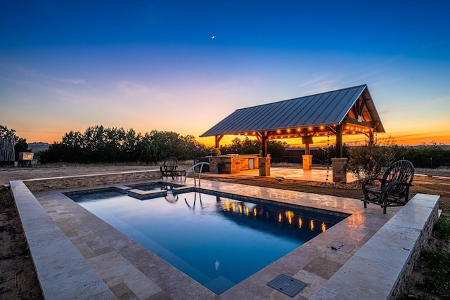 pool at dusk featuring a gazebo, exterior kitchen, an in ground hot tub, and a patio area