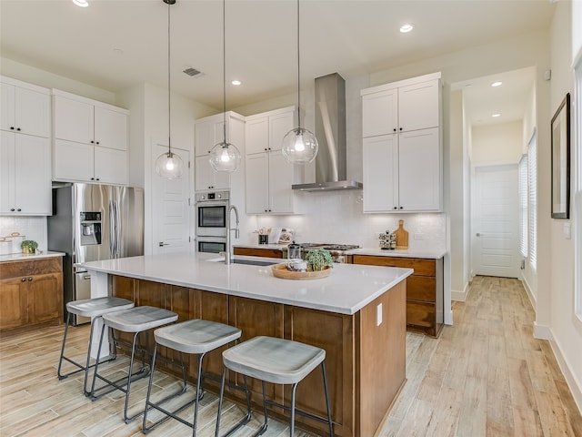 kitchen with white cabinets, light hardwood / wood-style floors, an island with sink, and wall chimney exhaust hood