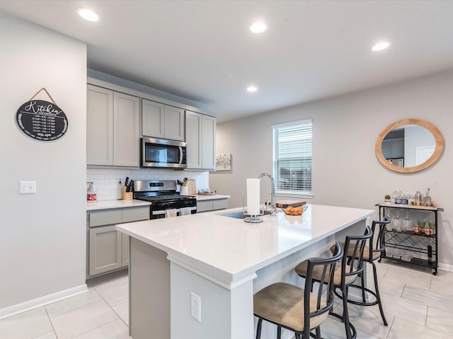 kitchen featuring a kitchen island with sink, a sink, appliances with stainless steel finishes, gray cabinets, and decorative backsplash