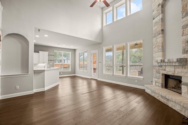 unfurnished living room with a stone fireplace, ceiling fan, dark hardwood / wood-style floors, a high ceiling, and sink