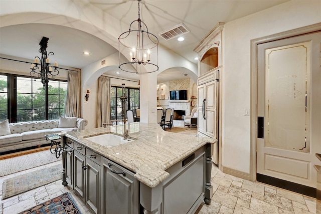 kitchen with gray cabinetry, decorative light fixtures, a chandelier, light stone countertops, and a kitchen island with sink