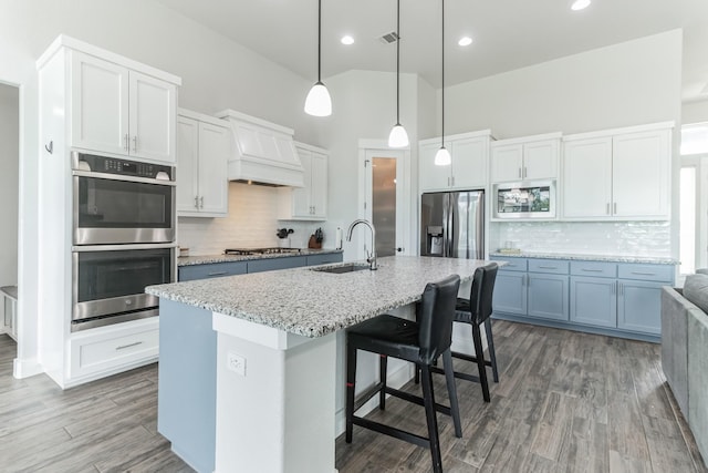 kitchen featuring a center island with sink, stainless steel appliances, custom range hood, sink, and white cabinetry