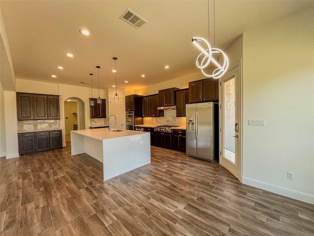 kitchen with a center island with sink, stainless steel appliances, dark brown cabinetry, decorative light fixtures, and backsplash