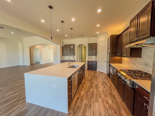 kitchen with stainless steel appliances, backsplash, dark brown cabinets, a center island with sink, and pendant lighting