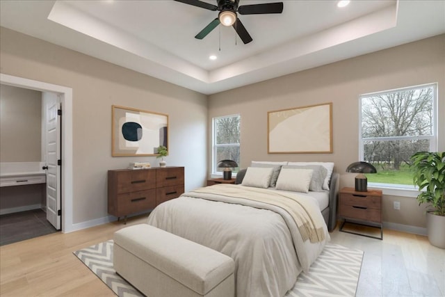 bedroom with baseboards, ensuite bathroom, recessed lighting, a tray ceiling, and light wood finished floors