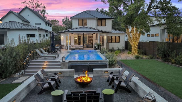 back of property at dusk featuring a fenced in pool, an outdoor living space with a fire pit, a standing seam roof, a patio area, and metal roof