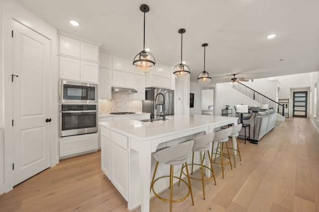 kitchen with stainless steel appliances, white cabinets, a center island with sink, light wood-type flooring, and backsplash