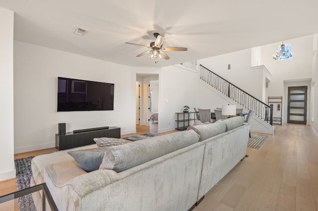 living room featuring baseboards, light wood finished floors, visible vents, ceiling fan with notable chandelier, and stairs