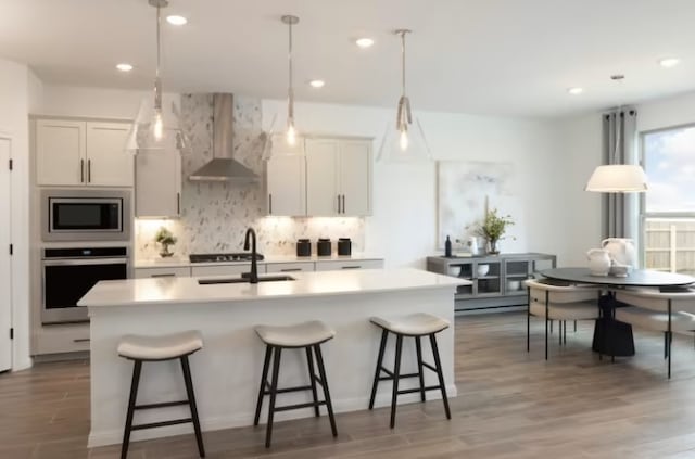 kitchen featuring appliances with stainless steel finishes, a kitchen island with sink, wall chimney range hood, white cabinets, and sink