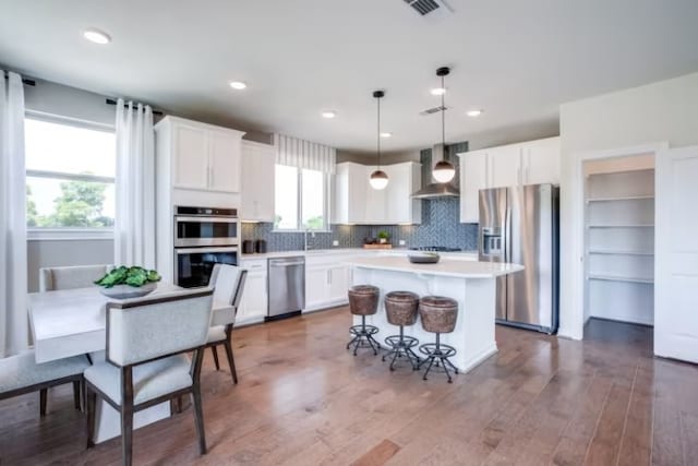 kitchen featuring appliances with stainless steel finishes, white cabinetry, wall chimney range hood, and decorative light fixtures