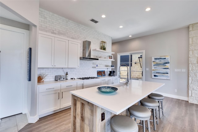 kitchen with wall chimney exhaust hood, a kitchen island with sink, backsplash, a breakfast bar area, and white cabinets