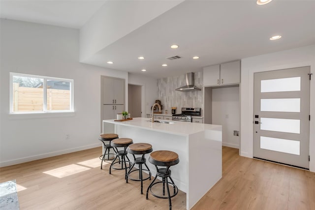 kitchen with stainless steel gas stove, an island with sink, light wood-type flooring, sink, and wall chimney range hood