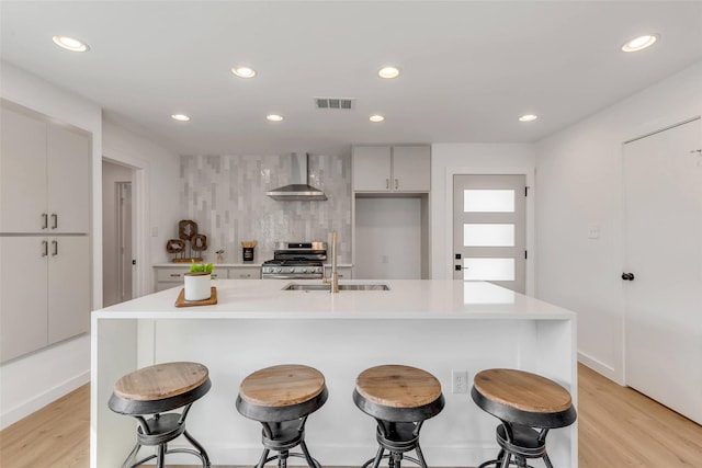 kitchen featuring sink, stainless steel range oven, wall chimney exhaust hood, light wood-type flooring, and a kitchen island with sink