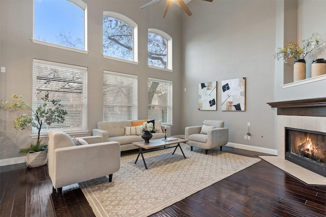 living room featuring ceiling fan, a tile fireplace, a towering ceiling, hardwood / wood-style flooring, and plenty of natural light