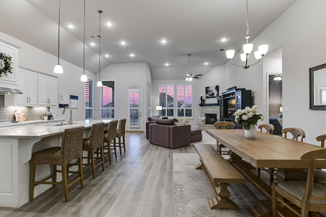 dining area with sink, high vaulted ceiling, light wood-type flooring, ceiling fan with notable chandelier, and a stone fireplace