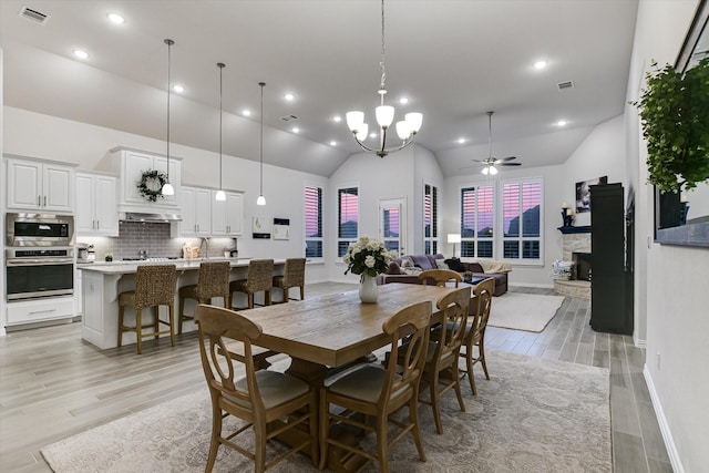 dining space featuring light wood-type flooring, high vaulted ceiling, ceiling fan with notable chandelier, and a fireplace