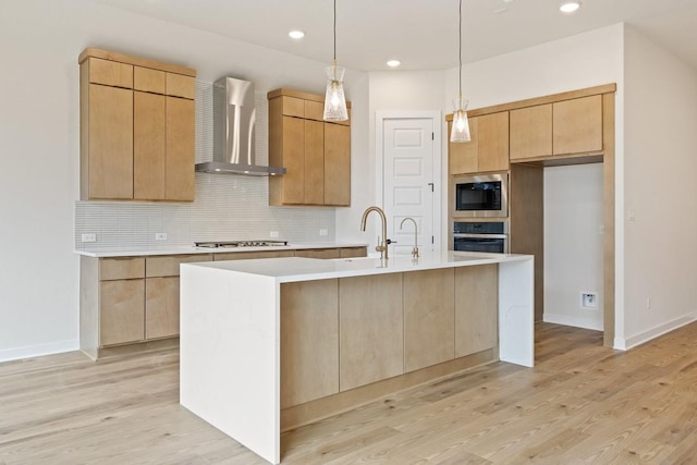 kitchen with appliances with stainless steel finishes, an island with sink, light brown cabinets, light hardwood / wood-style flooring, and wall chimney range hood