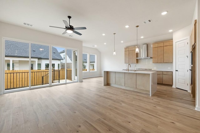 kitchen featuring light hardwood / wood-style floors, an island with sink, decorative backsplash, hanging light fixtures, and wall chimney range hood