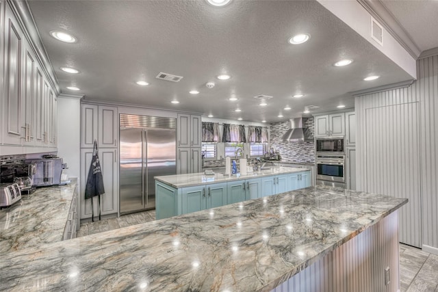 kitchen featuring a kitchen island with sink, built in appliances, kitchen peninsula, light stone counters, and wall chimney range hood