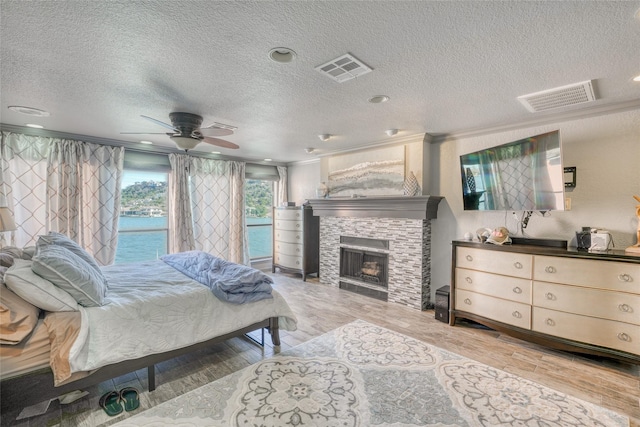 bedroom featuring a textured ceiling, ceiling fan, ornamental molding, light hardwood / wood-style flooring, and a stone fireplace