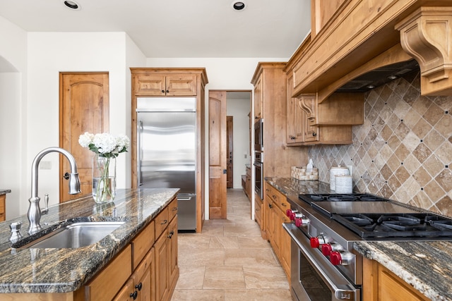 kitchen with high end appliances, custom range hood, backsplash, dark stone counters, and sink