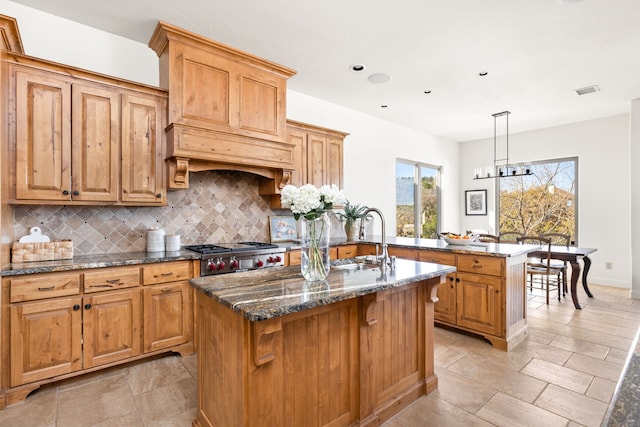 kitchen with hanging light fixtures, an island with sink, decorative backsplash, dark stone counters, and sink