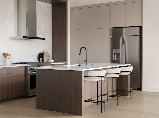 kitchen with stainless steel appliances, wall chimney exhaust hood, a kitchen island with sink, light hardwood / wood-style floors, and a breakfast bar