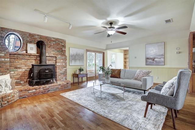 living room with wood-type flooring, a wood stove, track lighting, built in features, and ceiling fan