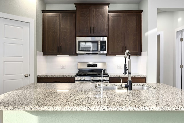 kitchen featuring light stone counters, a sink, stainless steel appliances, dark brown cabinetry, and tasteful backsplash