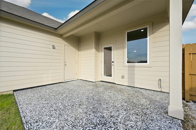 view of exterior entry featuring a shingled roof, a patio area, and fence