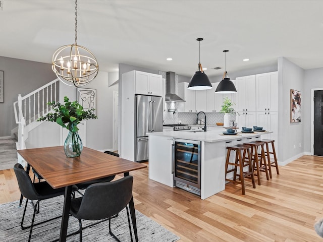 kitchen with an island with sink, appliances with stainless steel finishes, wall chimney range hood, pendant lighting, and white cabinets