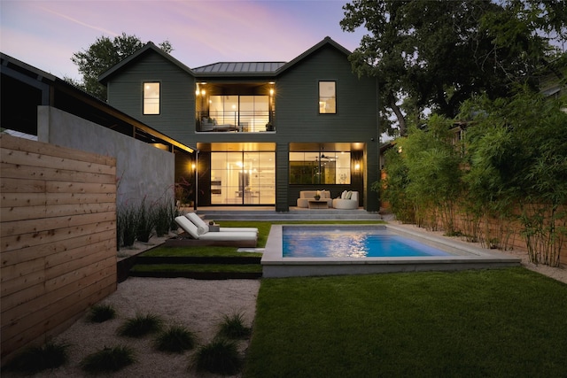 back house at dusk featuring a balcony, outdoor lounge area, and a lawn