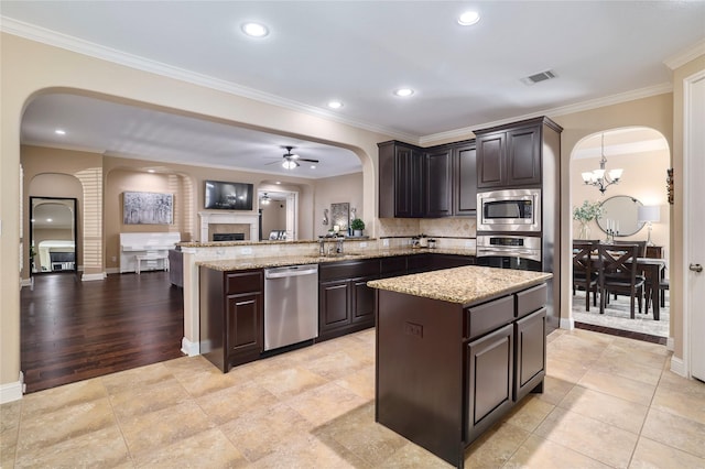 kitchen featuring crown molding, dark brown cabinetry, a tiled fireplace, appliances with stainless steel finishes, and ceiling fan with notable chandelier